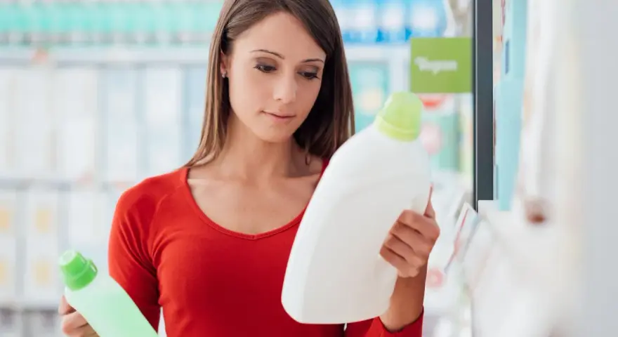 Mujer compara dos botellas de detergente, leyendo la etiqueta producto de limpieza.