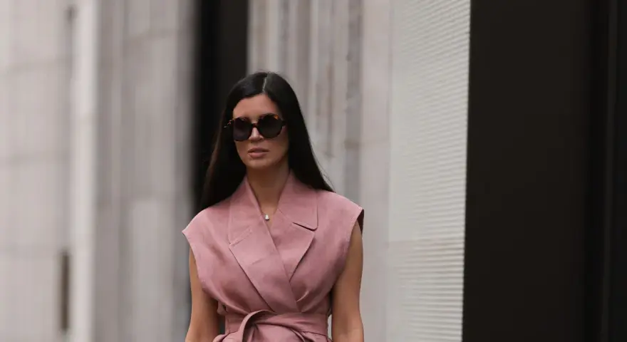 Mujer con gafas de sol y un elegante chaleco de lino rosa, atado a la cintura, pasea por la ciudad.