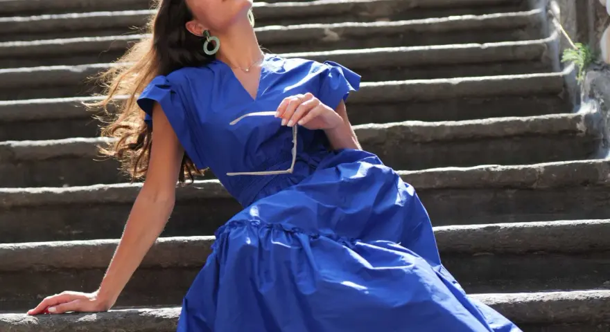 Mujer con vestido azul marino, gafas de sol en mano, sentada en escalones de piedra. Perfecta para combinar con zapatos para vestido azul marino.