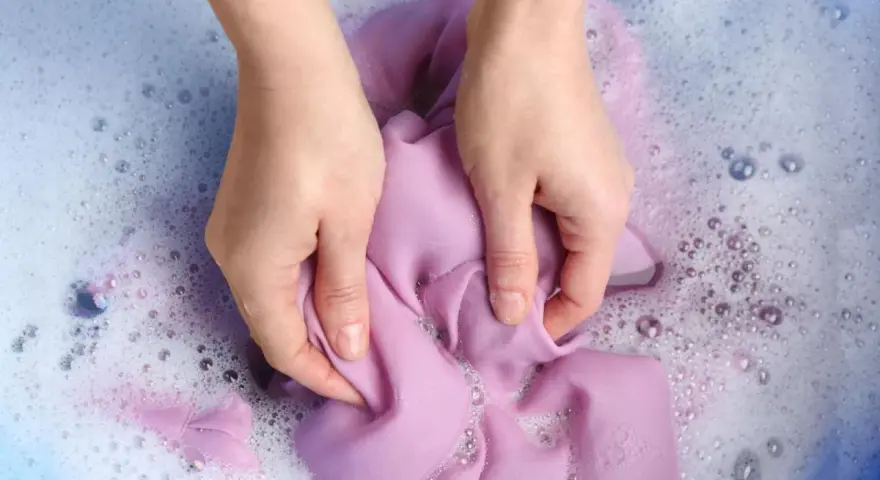 Manos lavando ropa morada a mano en agua con jabón y burbujas.