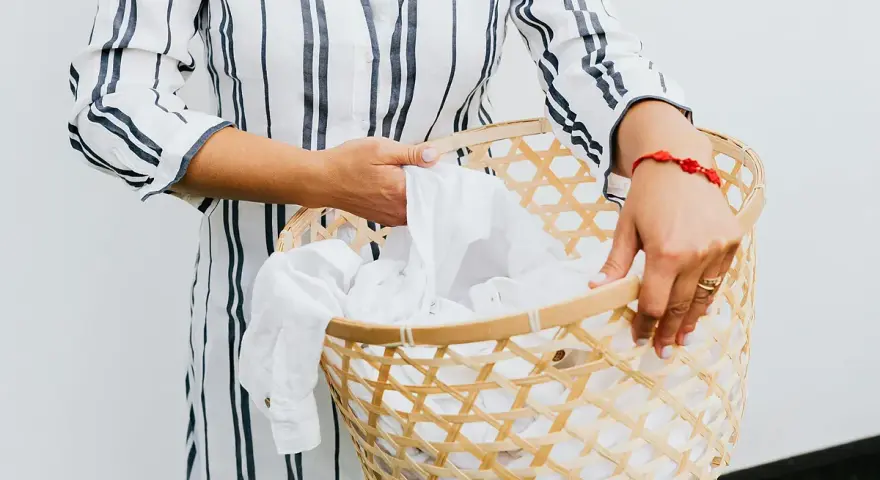 Manos sostienen una cesta de mimbre llena de ropa blanca, lista para lavar camiseta a mano.