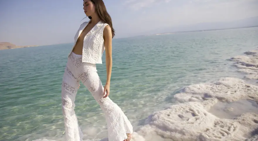 Mujer con traje blanco de encaje, posando en la orilla de un mar turquesa con formaciones de sal. Un look ibiza perfecto para el verano.