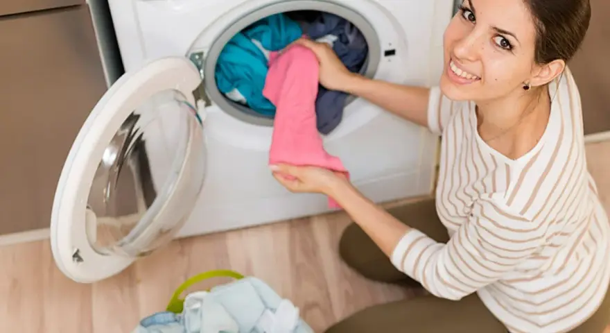 Mujer sonriente carga ropa en lavadora. El detergente sirve para dejar la ropa impecable y eliminar manchas.
