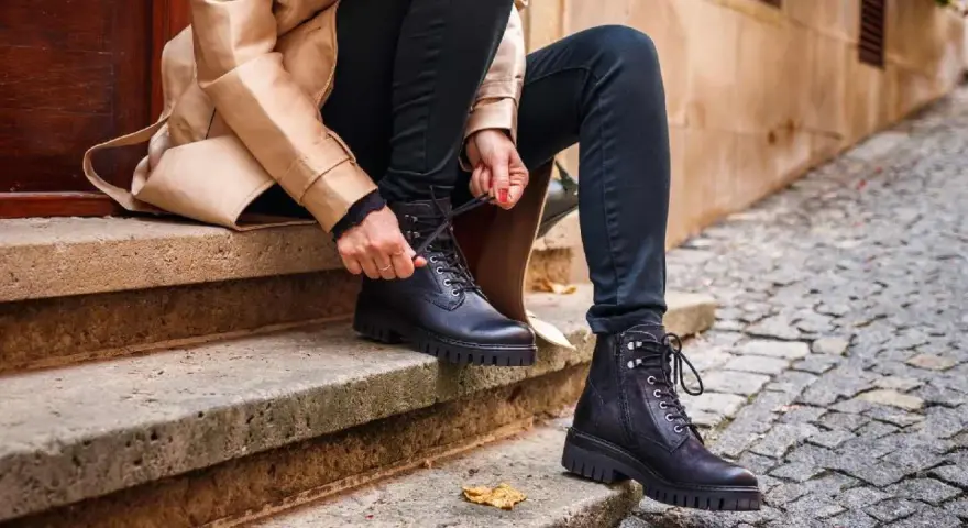 Persona se sienta en escalones de piedra, atándose unos pantalones anchos con botas militares negras.