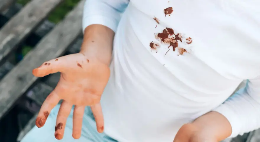 Manos y camiseta blanca con manchas de chocolate. Un niño muestra sus manos sucias y su ropa manchada, buscando cómo quitar manchas de chocolate ya lavadas.