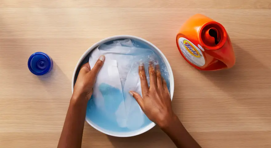 Manos lavando ropa blanca en un bol con agua y detergente. A un lado, un bote de detergente y un tapón azul.