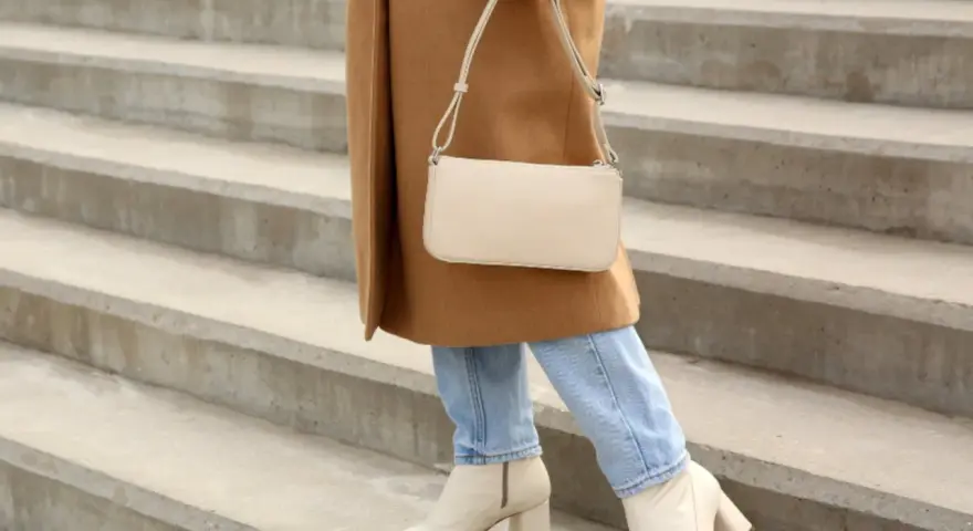 Mujer con abrigo camel, jeans y bolso crema, lista para combinar outfit con botas beige.