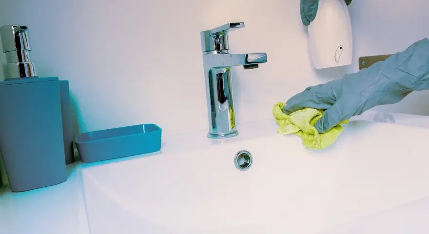 Manos con guantes limpian lavabo. Se usa un paño amarillo y un dispensador blanco.