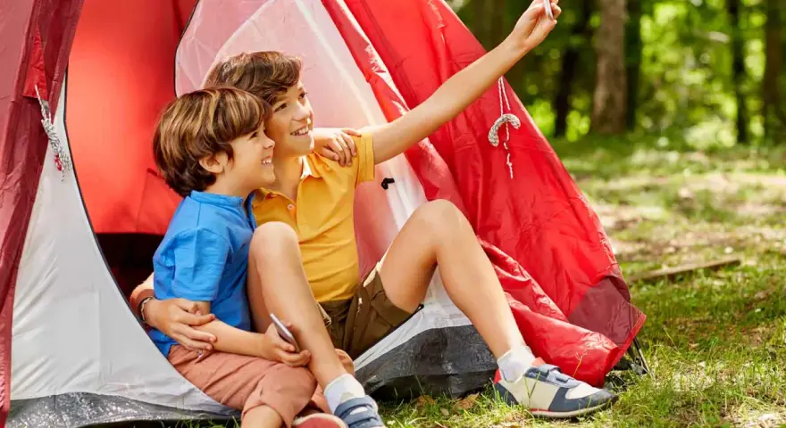Dos niños, uno con talla niño 7 años, se toman un selfie sonrientes dentro de una tienda de campaña roja en un bosque.