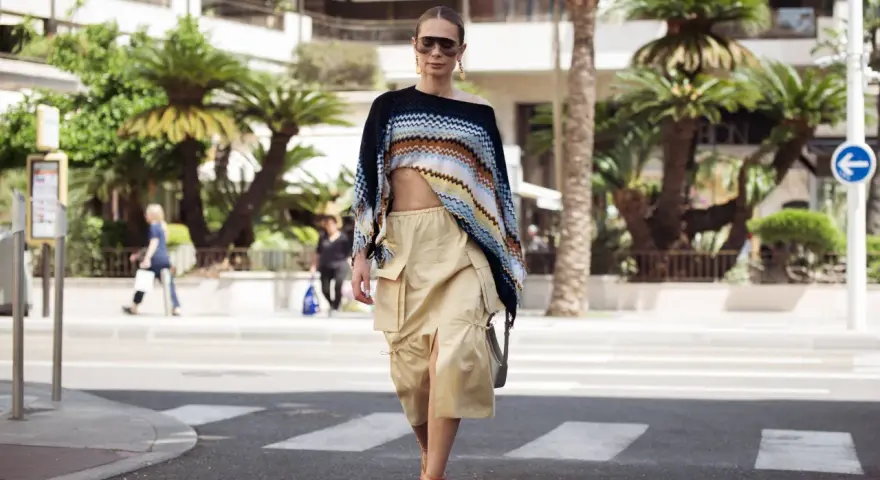 Mujer con un top corto de crochet y una falda vaporosa color beige, paseando por la calle con palmeras de fondo.