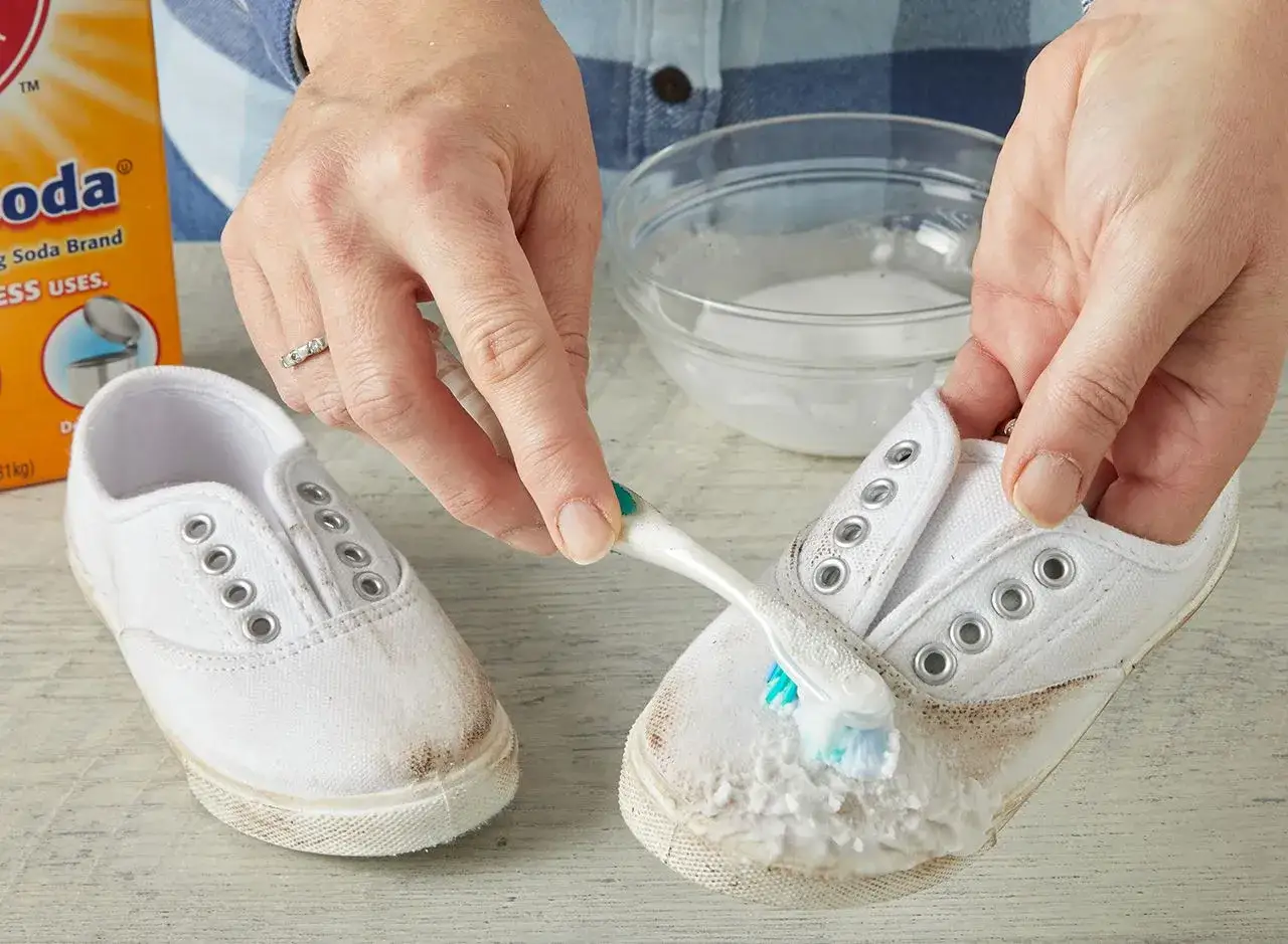 Manos limpian botines blancos con cepillo y bicarbonato.