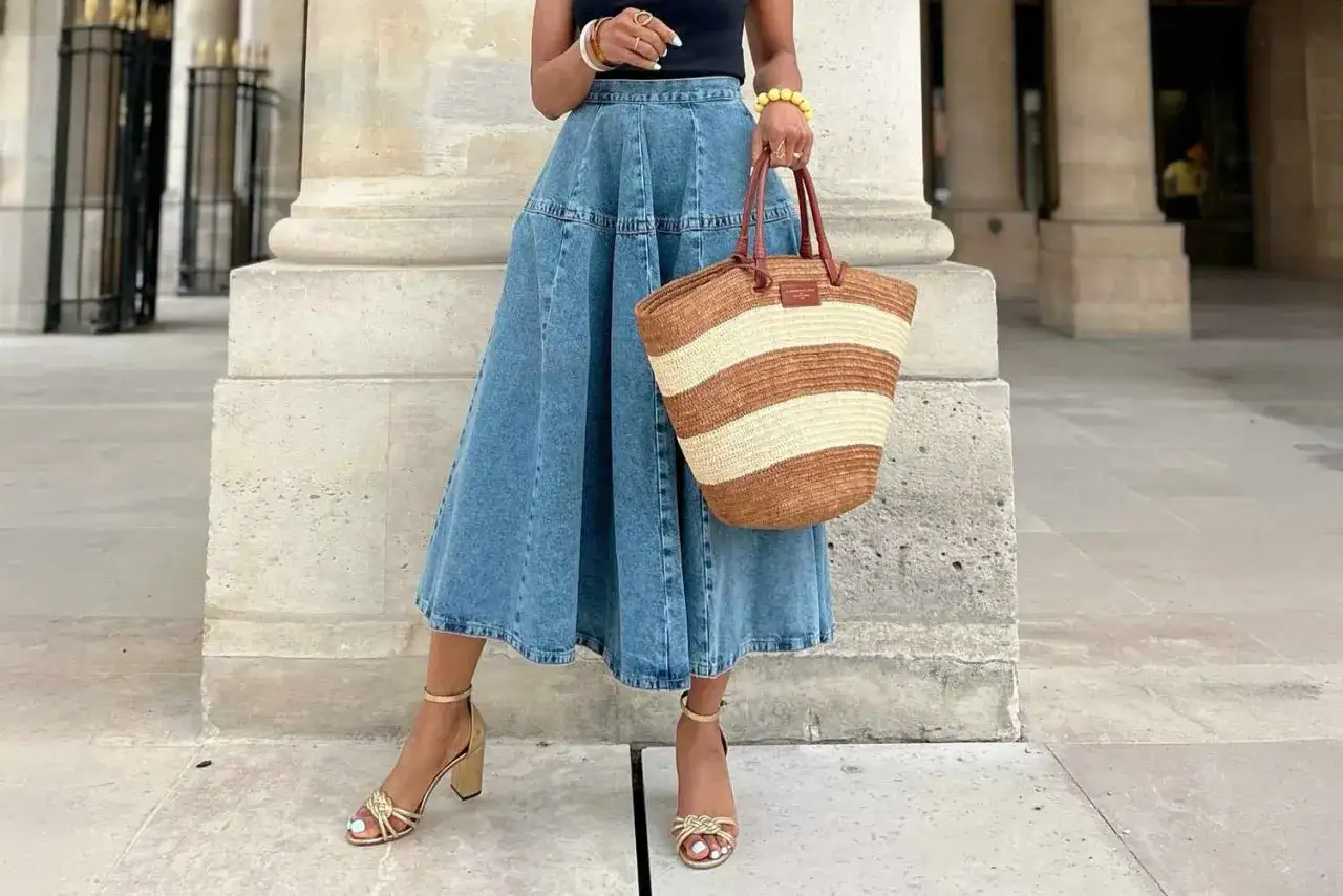 Una mujer luce una falda vaporosa vaquera, sandalias doradas y un bolso de mimbre a rayas, lista para un paseo veraniego.