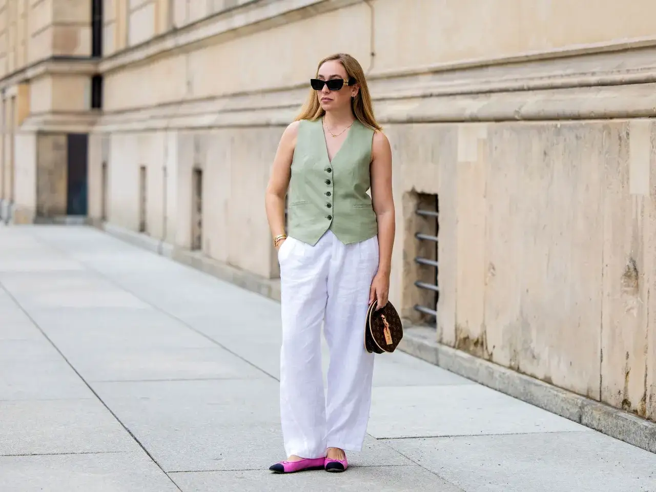 Mujer con chaleco verde y pantal&oacute;n lino blanco, gafas de sol y bolso marr&oacute;n.