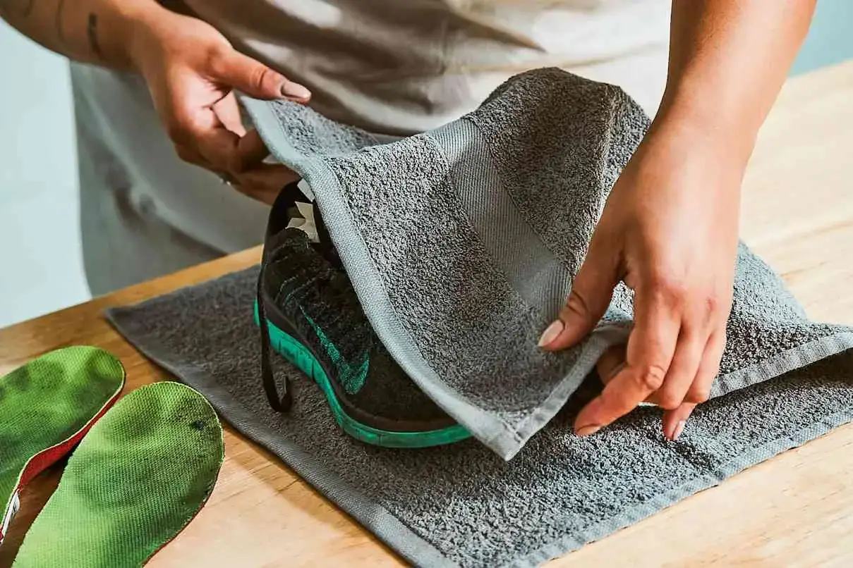 Manos secan zapatillas deportivas negras con detalles verdes con una toalla gris. Se pueden meter las zapatillas en la secadora si se secan bien.