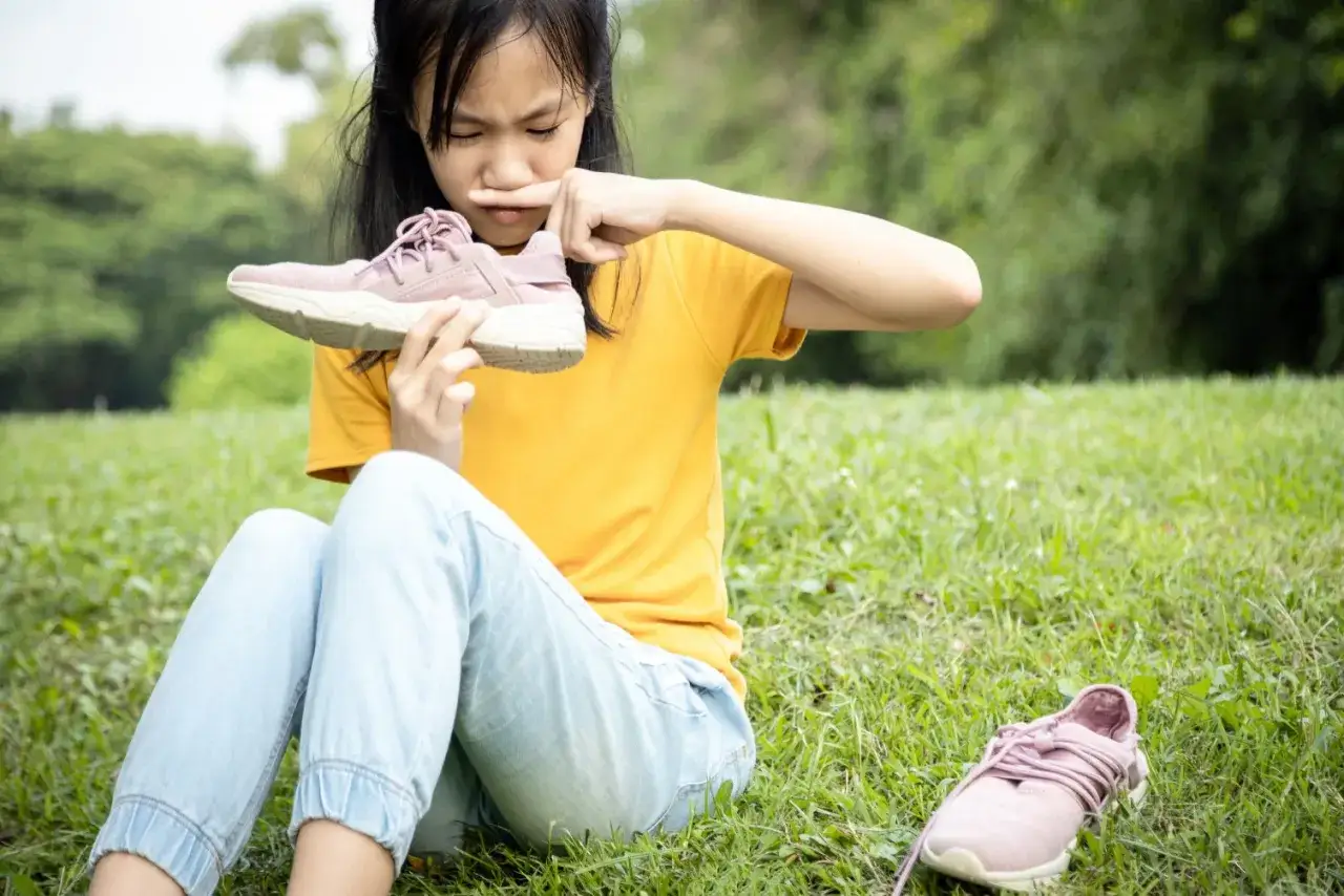 Ni&ntilde;a con expresi&oacute;n de asco huele zapatilla, quiz&aacute;s por el sudor de los pies de ni&ntilde;os tras un d&iacute;a de juegos.