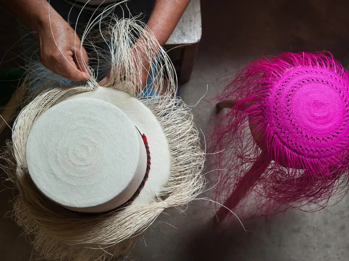 Manos tejiendo un sombrero blanco con hebras de palma de iraca. A su lado, otro sombrero rosa vibrante con flecos.