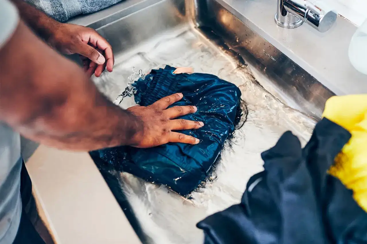 Man's hands gently wash a dark blue garment in a sink, demonstrating how to hand wash clothes.