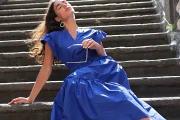 Mujer con vestido azul marino, gafas de sol en mano, sentada en escalones de piedra. Perfecta para combinar con zapatos para vestido azul marino.