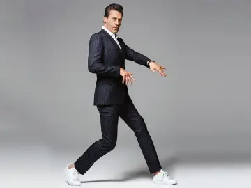 Hombre con traje a rayas y zapatillas deportivas blancas, en pose dinámica sobre fondo gris.