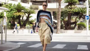 Mujer con un top corto de crochet y una falda vaporosa color beige, paseando por la calle con palmeras de fondo.