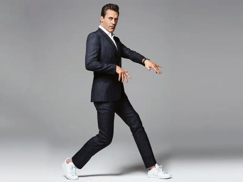 Hombre con traje a rayas y zapatillas deportivas blancas, en pose dinámica sobre fondo gris.